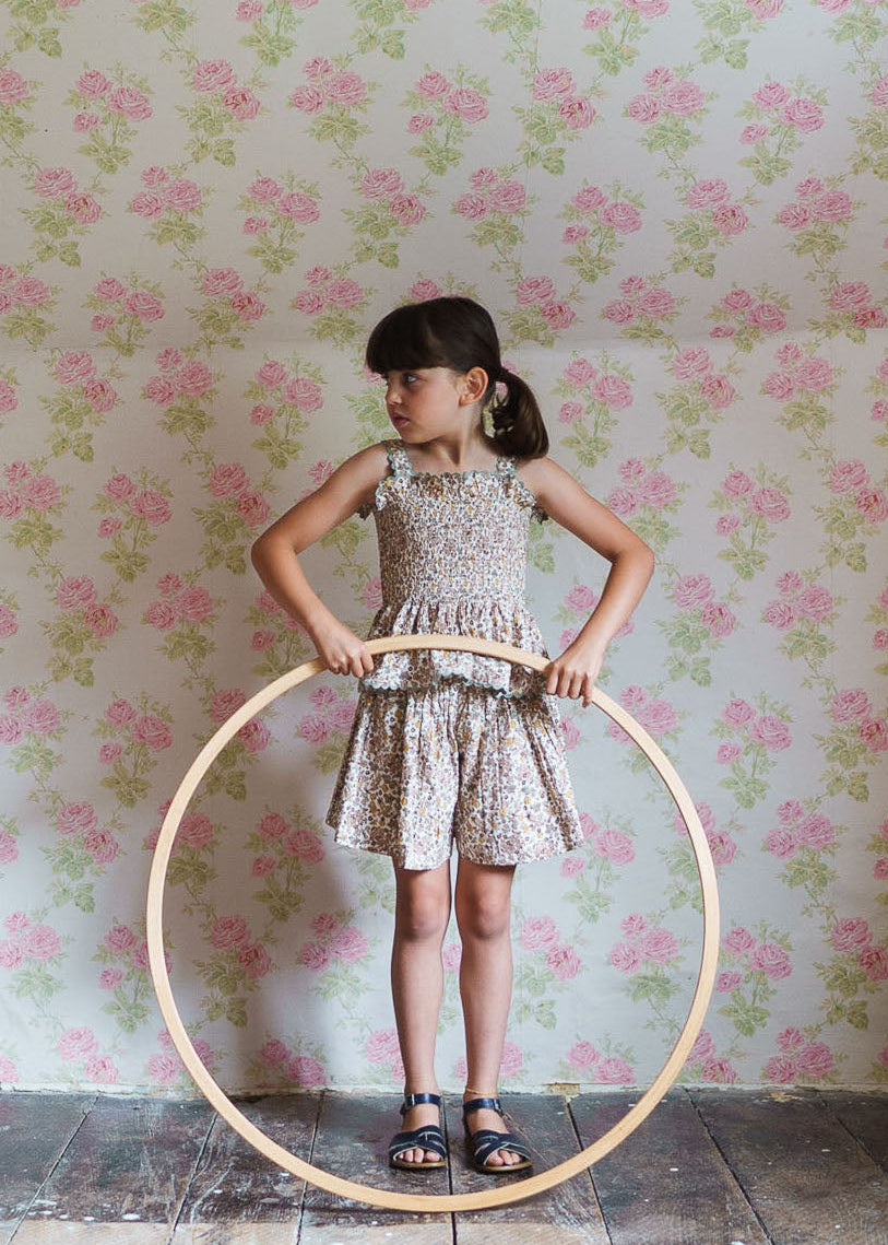 Young girl wearing organic cotton floral print culottes in a vintage style, standing inside a wooden hoop against a floral wallpaper, embodying sustainable children's clothing.