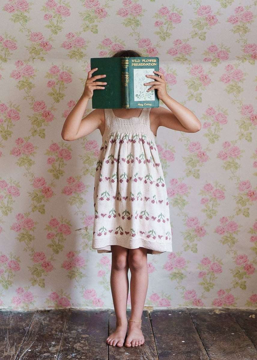 Child wearing a hand-knitted cotton pinafore dress with cherry pattern, standing barefoot on wooden floor against vintage floral wallpaper, holding a book in front of face.