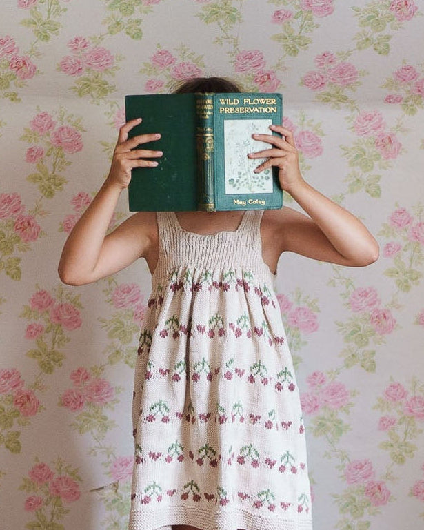 Child wearing a hand-knitted cotton pinafore dress with cherry pattern, standing barefoot on wooden floor against vintage floral wallpaper, holding a book in front of face.