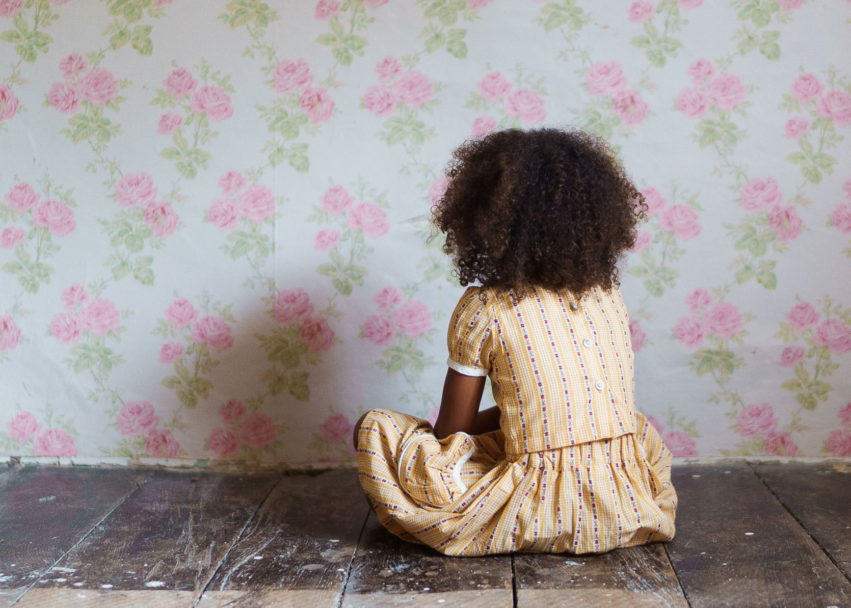 Child wearing a vintage style girls cotton top and skirt with a buttercup floral pattern, sitting on a rustic wooden floor against a floral wallpaper background.