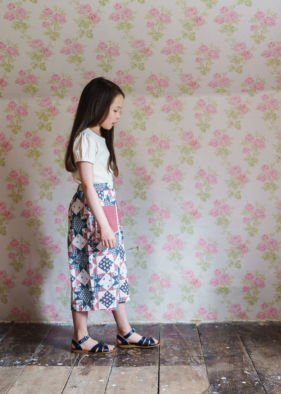 Young girl wearing vintage style patchwork floral blue organic cotton trousers, standing against a soft pink floral wallpaper, showcasing sustainable children's clothing.