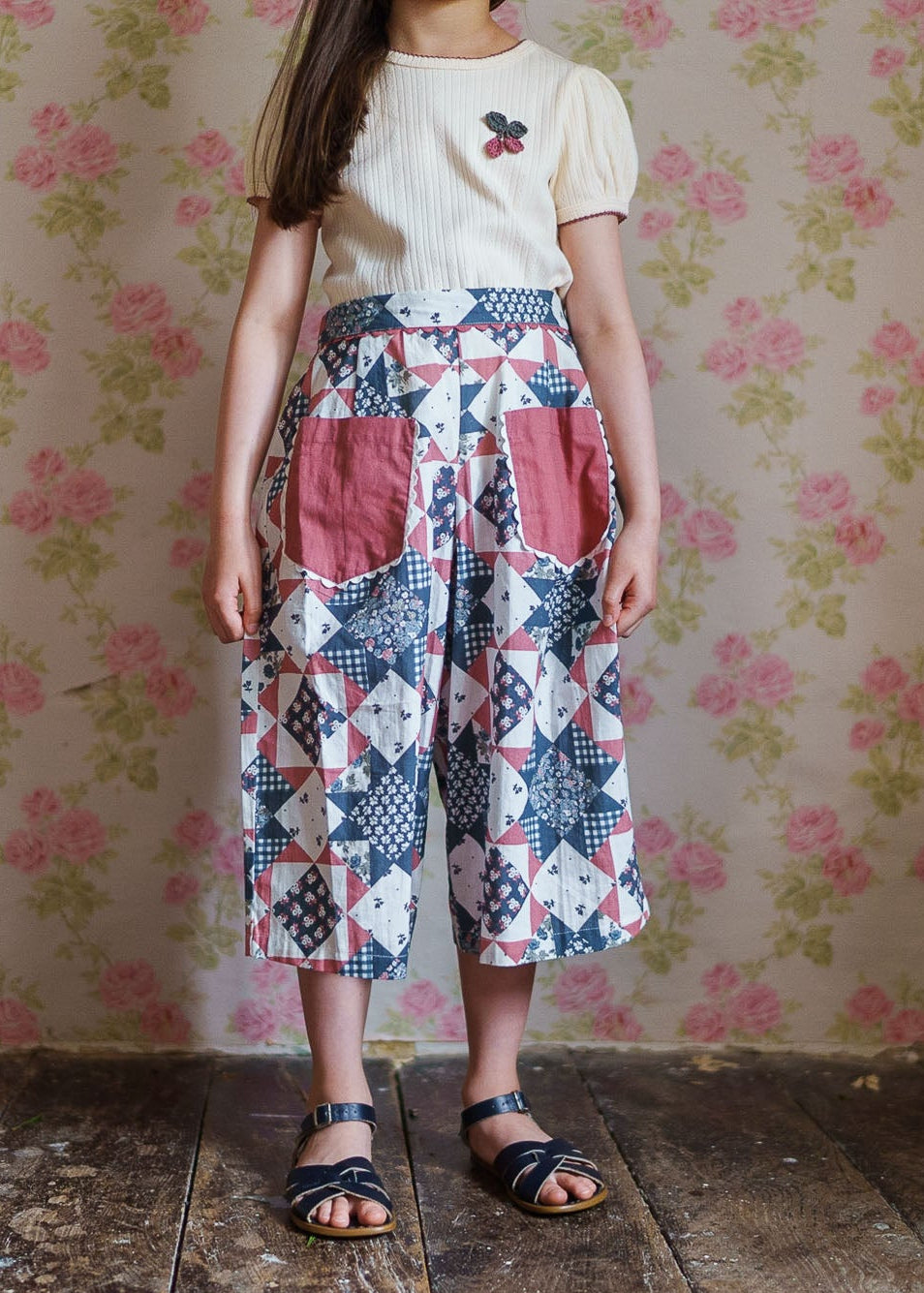 Child wearing vintage style patchwork floral organic cotton trousers with large pink pockets, paired with a cream short-sleeve top, standing against a floral wallpaper background.