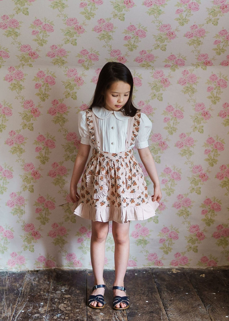 Young girl wearing a cream organic cotton blouse paired with a vintage style floral pinafore dress, standing against a floral patterned wall in a natural, sustainable children’s clothing setting.