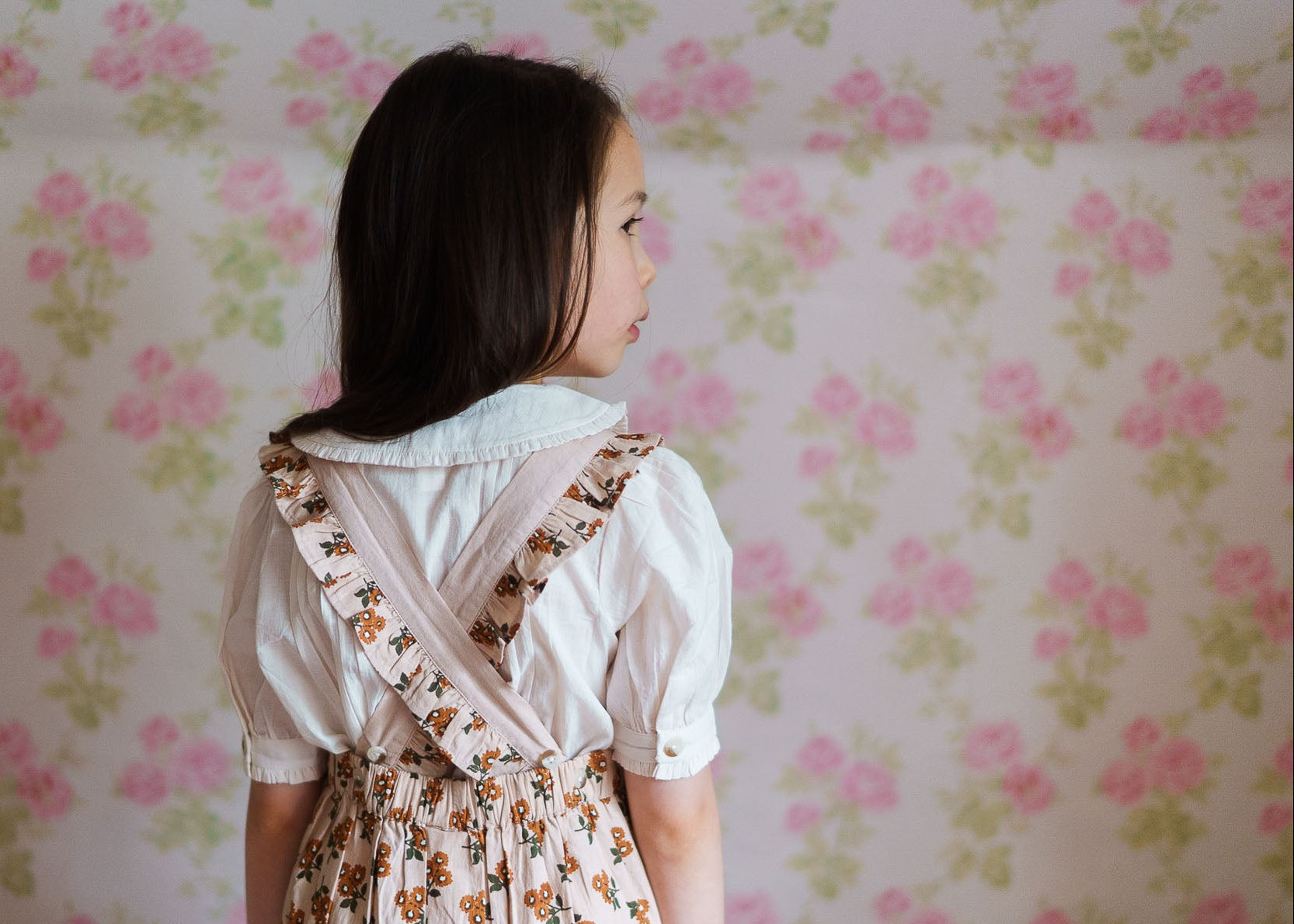 Young girl wearing a cream organic cotton blouse with vintage-style floral pinafore, standing against a softly patterned floral wallpaper background in a warm, natural setting.