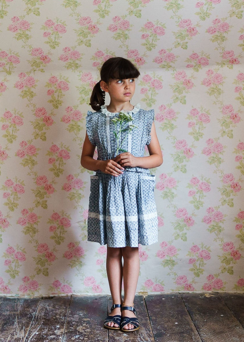 Young girl wearing a sustainable blue floral vintage-style dress made from organic cotton, standing against a pink rose patterned wall holding a small flower.