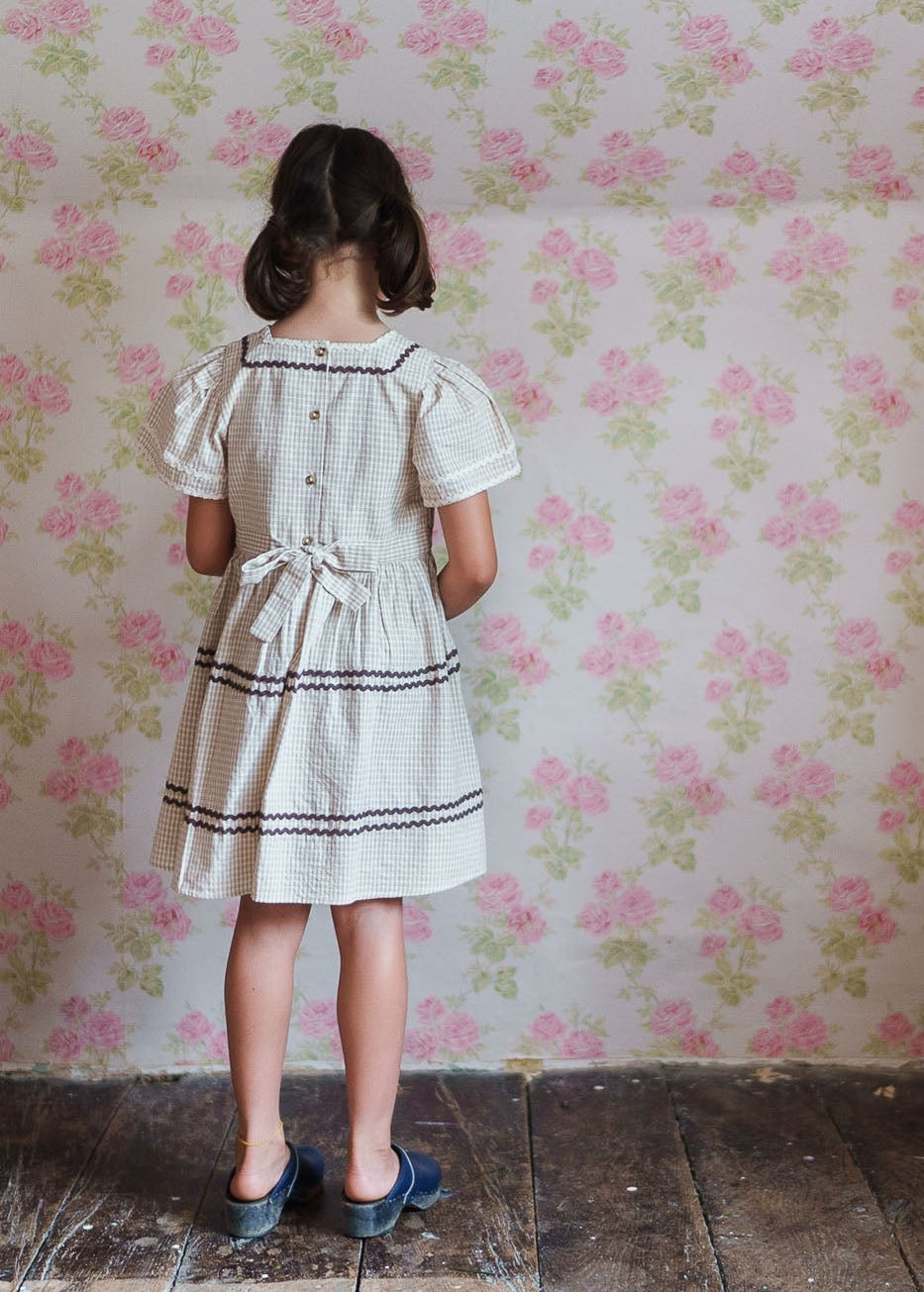 Young girl wearing a vintage-style yellow check cotton blend dress with puffed sleeves and smocked detailing, standing against floral wallpaper in a natural, ethical children's clothing setting.