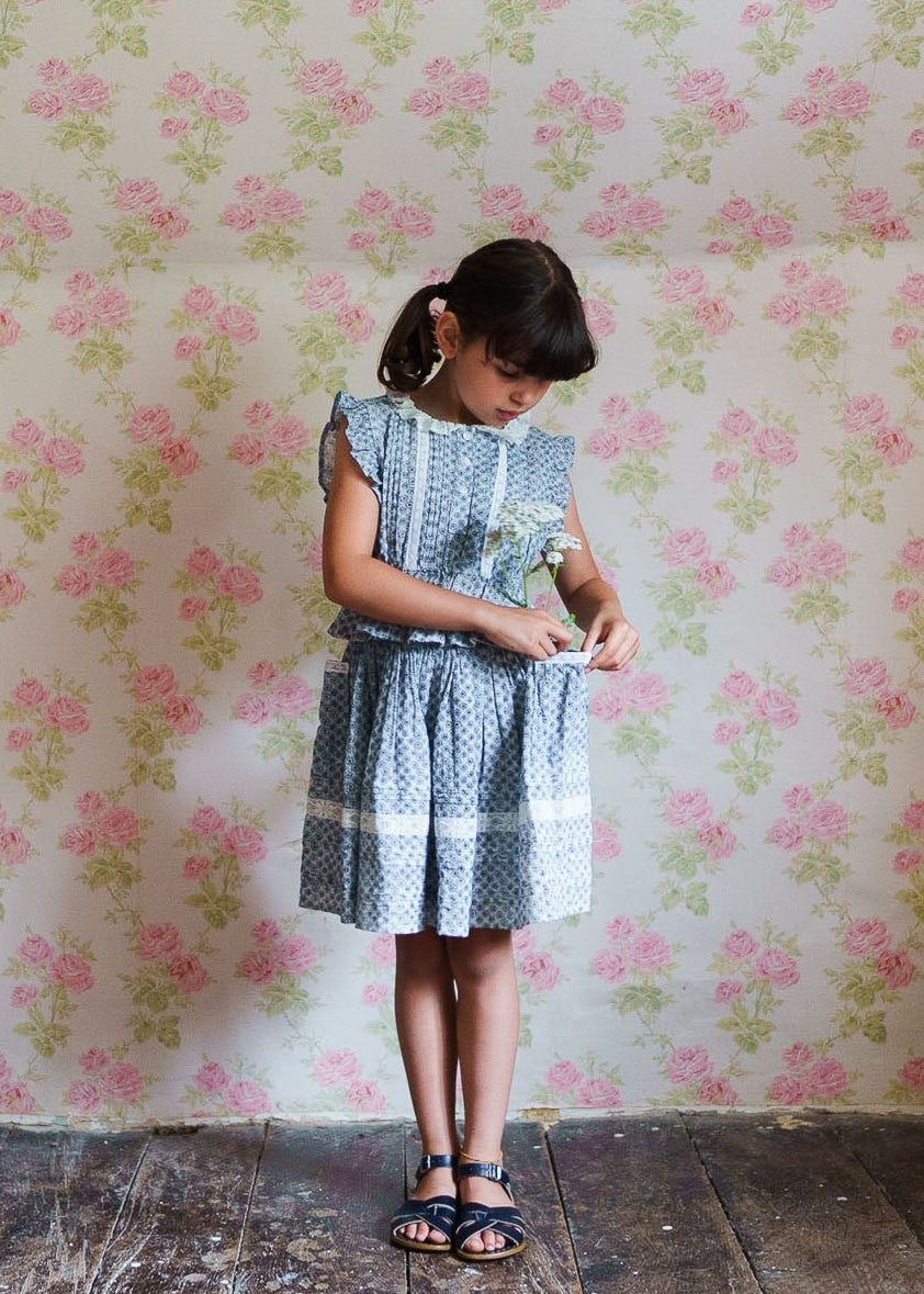 Young girl in vintage style blue floral dress made from organic cotton, standing against a floral wallpaper, showcasing ethically made sustainable children's clothing.