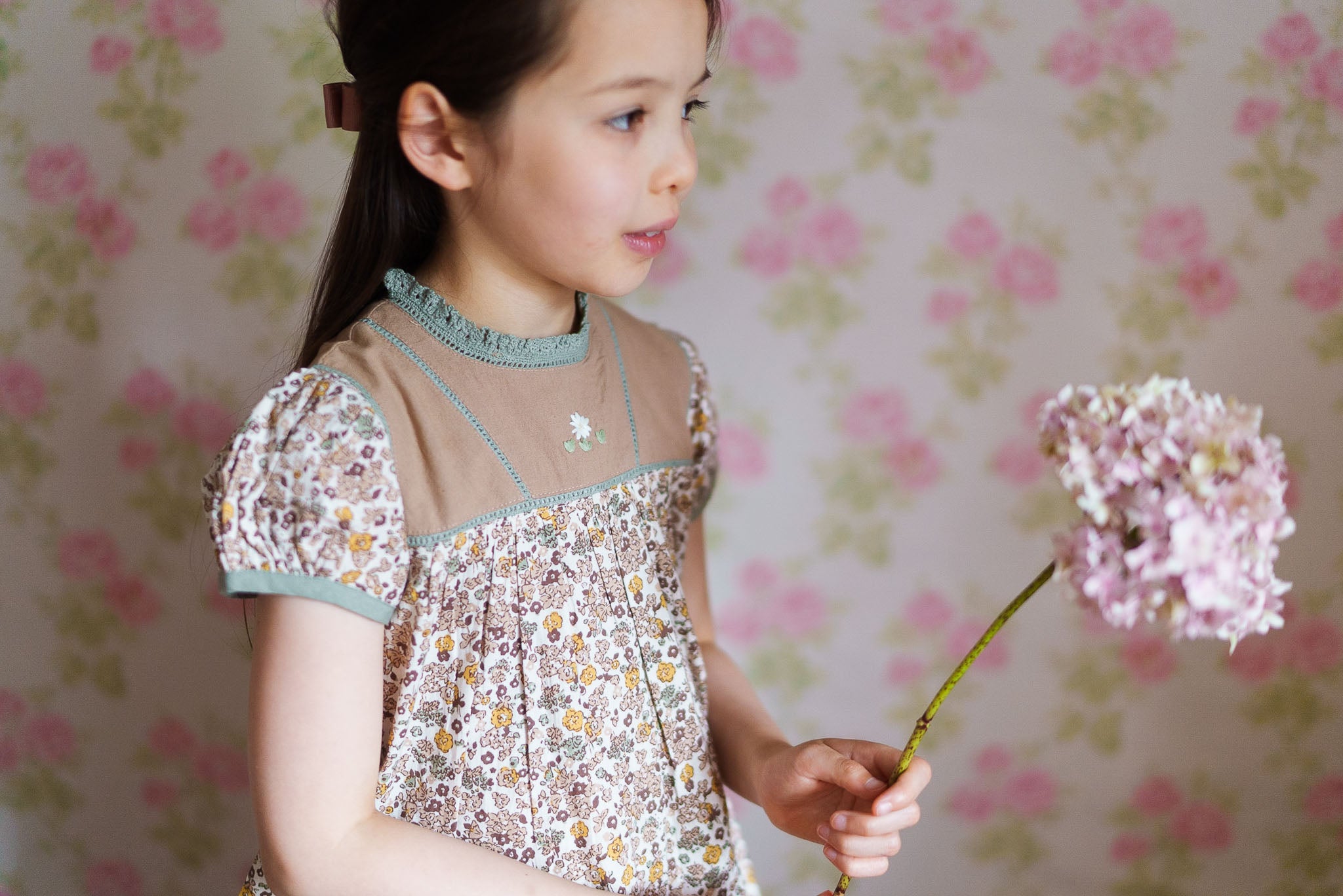 Young girl wearing a vintage style floral cotton blouse with delicate lace details, holding a pink flower, showcasing natural and ethically made children’s clothing.