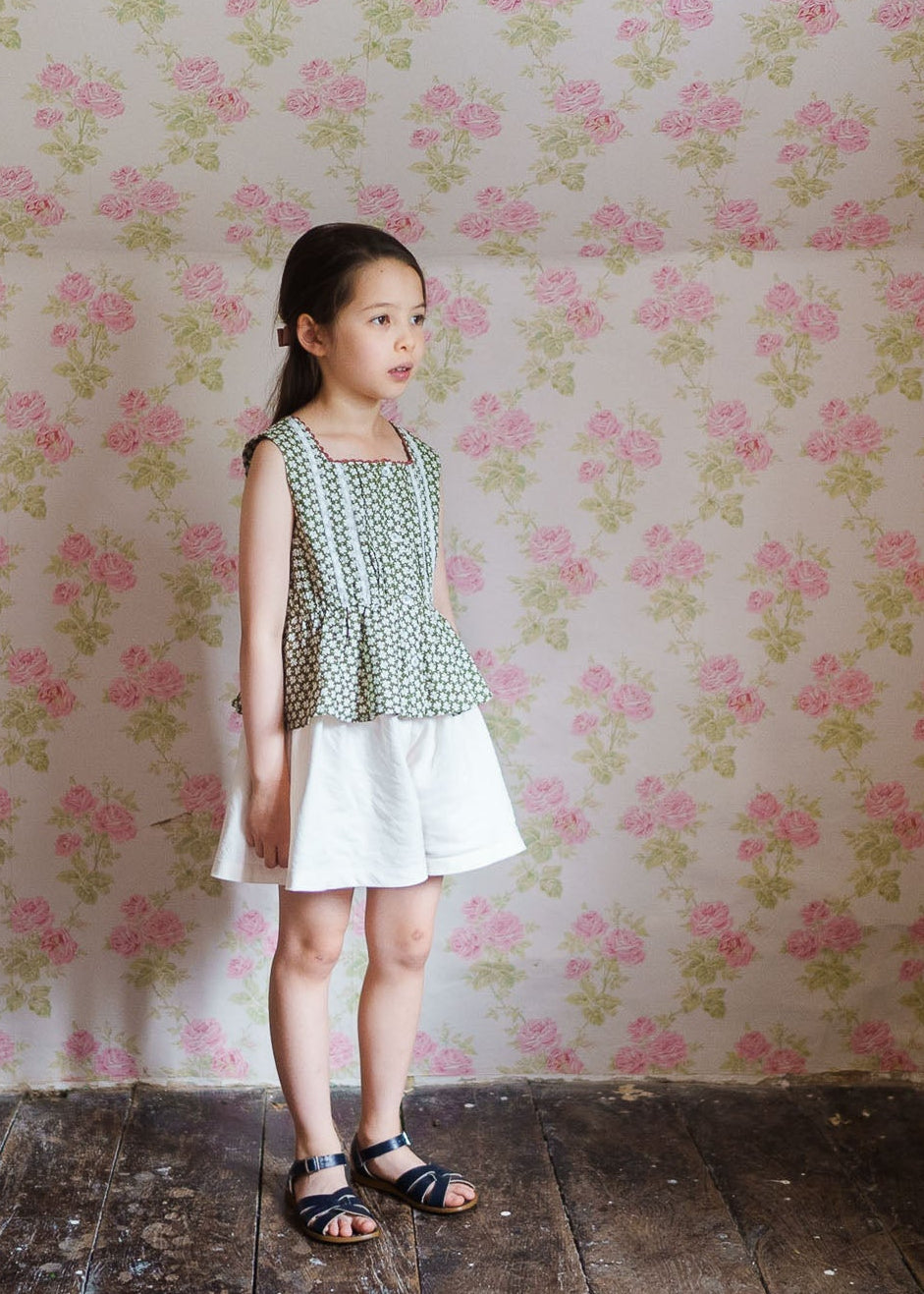 Young girl wearing a vintage style floral sun top and white skirt standing against a pink and green floral wallpaper, showcasing sustainable and organic children's clothing.