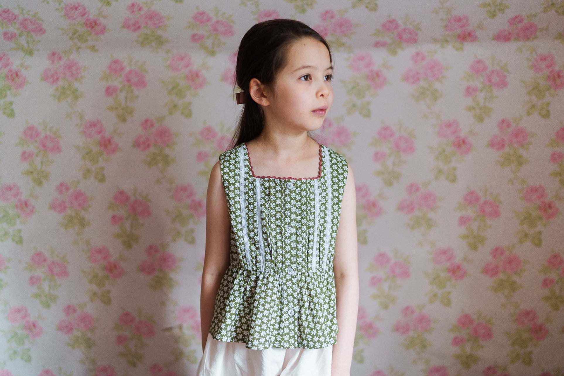 Young girl wearing a vintage-style green floral top made from organic cotton, standing in front of a soft pink and green floral wallpaper background.