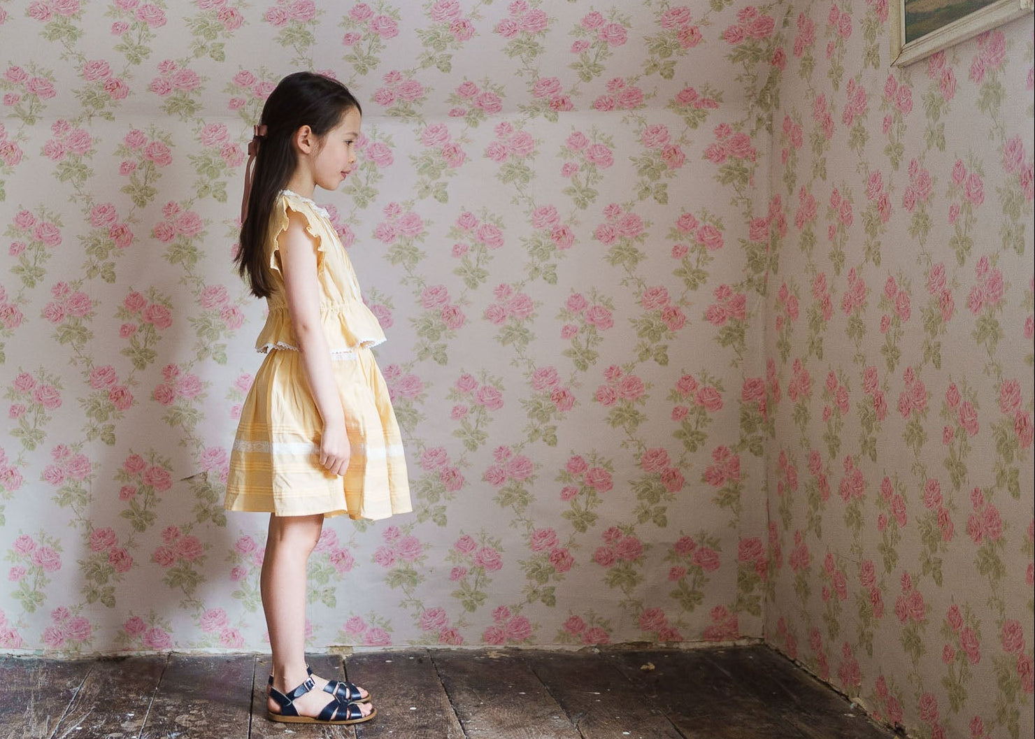 Young girl wearing a vintage style yellow organic cotton blouse and skirt standing in a room with floral wallpaper and wooden floors, reflecting sustainable and ethical children's clothing.