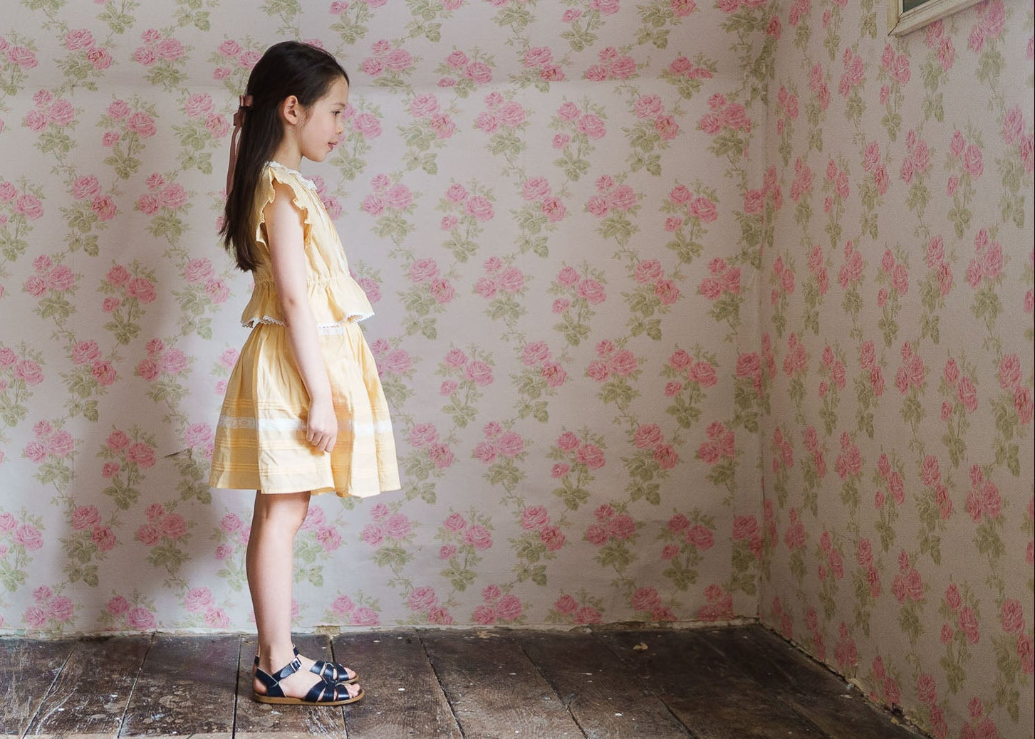 Young girl wearing a sustainable yellow organic cotton skirt with ruffled top, standing in a vintage-style room with floral wallpaper, showcasing ethical children's clothing.