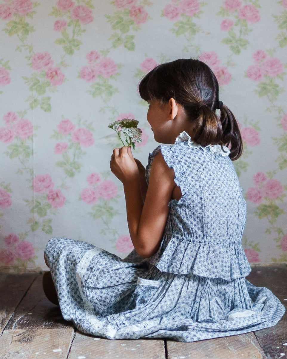 Young girl in a vintage-style blue floral blouse sitting on a rustic wooden floor, holding a small flower, surrounded by soft pink floral wallpaper, showcasing natural and organic children's clothing.