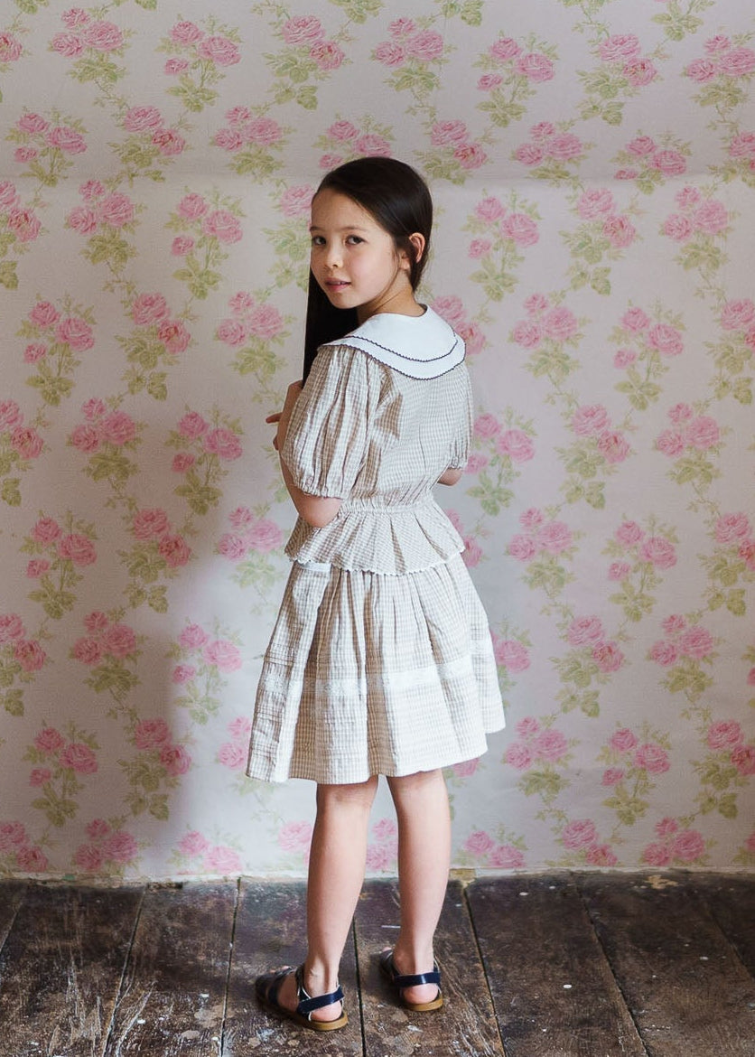 Young girl wearing a vintage-inspired straw check blouse with a white collar, standing in a softly lit room with floral wallpaper, showcasing natural, organic children’s clothing.
