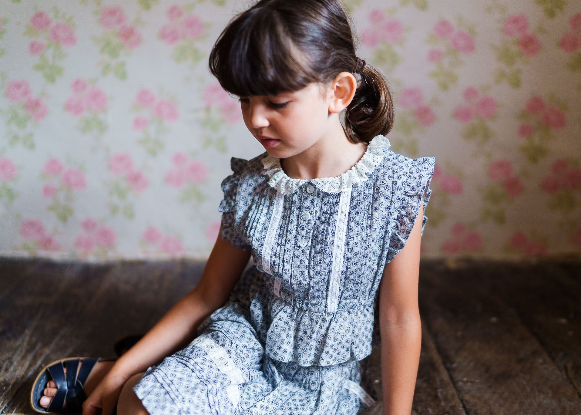 Young girl wearing a vintage-style girls blouse with delicate floral patterns and lace details, made from organic cotton in a softly lit room with floral wallpaper.