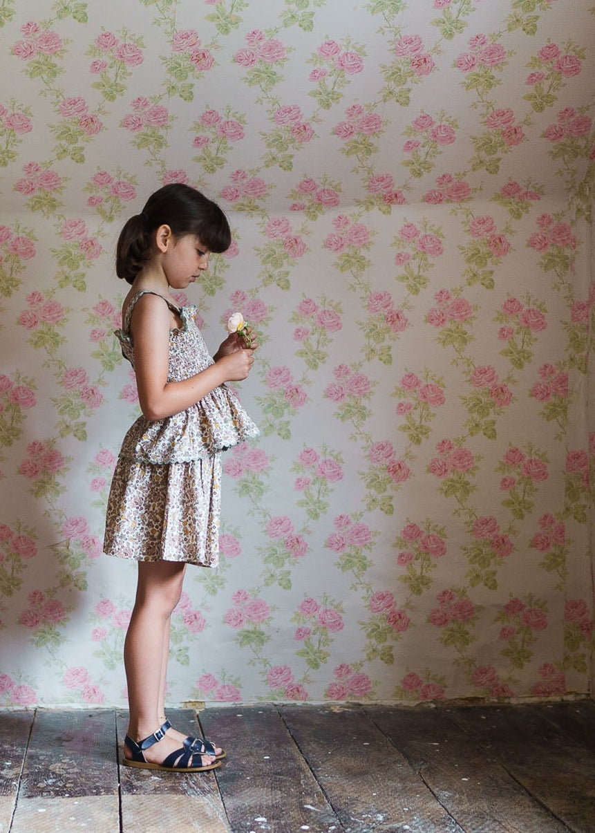 Young girl in vintage-style floral cotton dress holding a flower, standing against a softly patterned pink and green floral wallpaper in a rustic room.