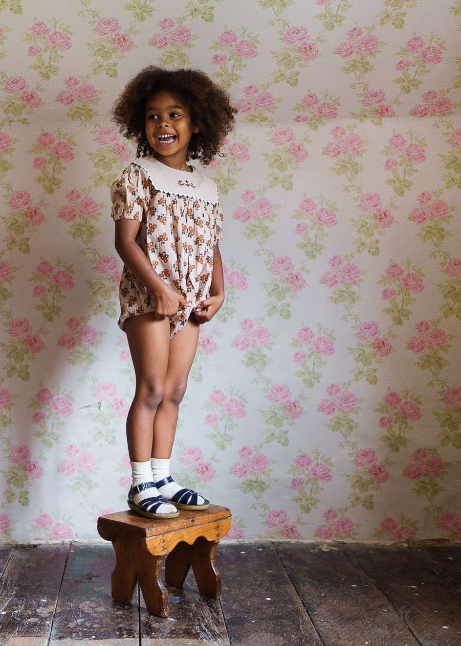 Happy young girl standing on a wooden stool wearing a vintage-style organic cotton romper with a floral pattern, showcasing sustainable and ethically made children's clothing.