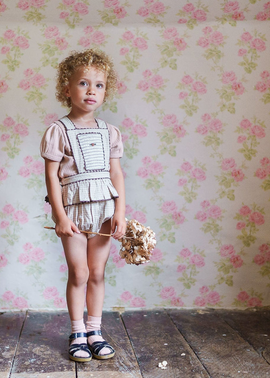 Young child with curly hair wearing a vintage-style cotton romper in straw check pattern, standing on a wooden floor against a floral wallpaper, holding dried flowers.