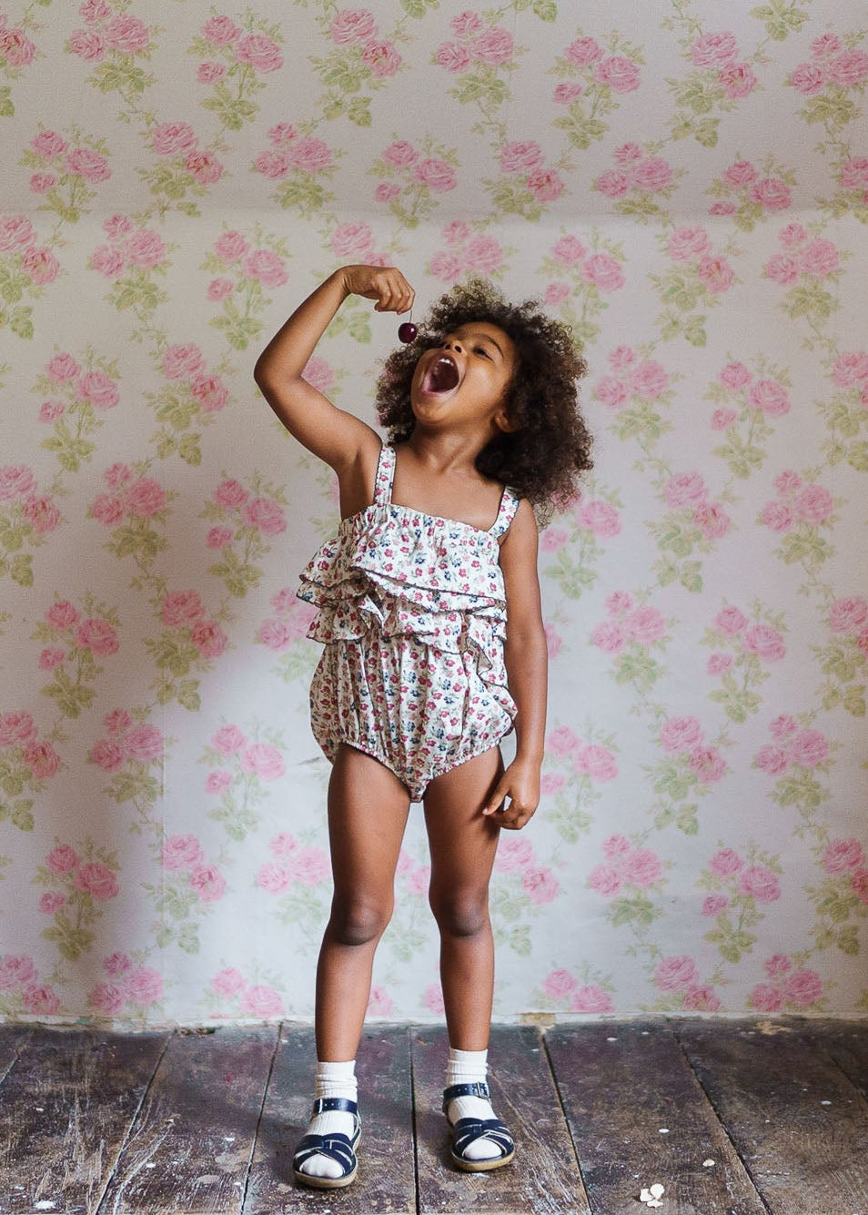 Young girl wearing a floral organic cotton romper with ruffles, standing on wooden floor against vintage-style floral wallpaper, joyfully holding a cherry above her open mouth.