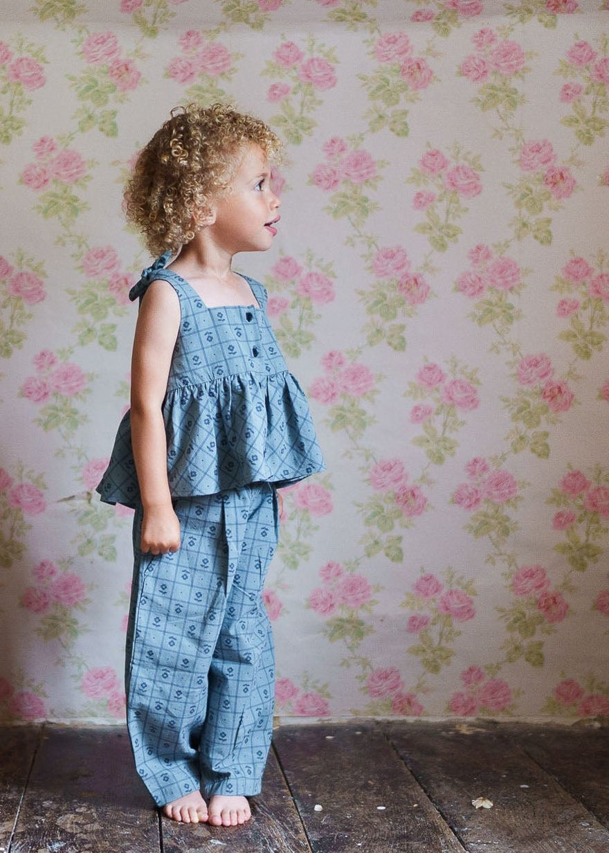 Child wearing vintage-style blue organic cotton trousers and matching top, standing barefoot on rustic wooden floor against floral wallpaper in a natural, ethical children's clothing setting.