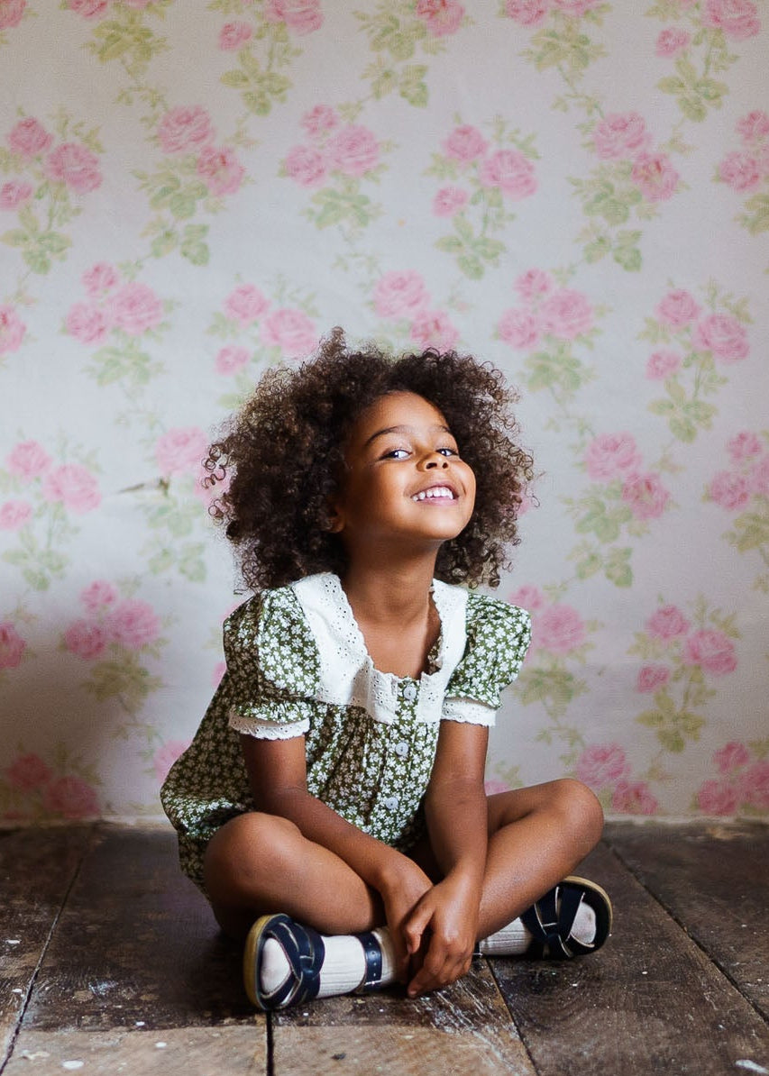 Smiling young girl sitting cross-legged on a wooden floor wearing a vintage style green floral romper with a white lace collar, against a soft floral wallpaper backdrop, showcasing sustainable, organic children's clothing.