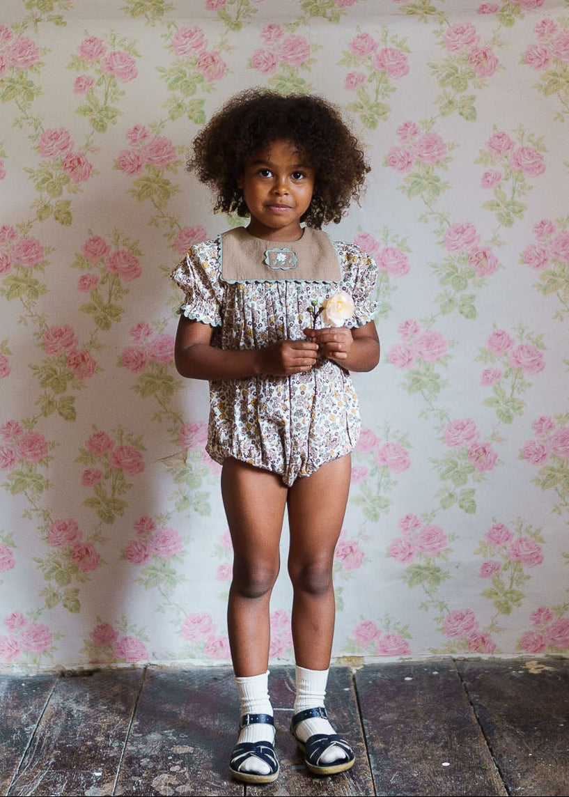 Young child wearing a vintage-style floral romper made from organic cotton, standing against a soft pink and green rose patterned wall, holding a small flower.