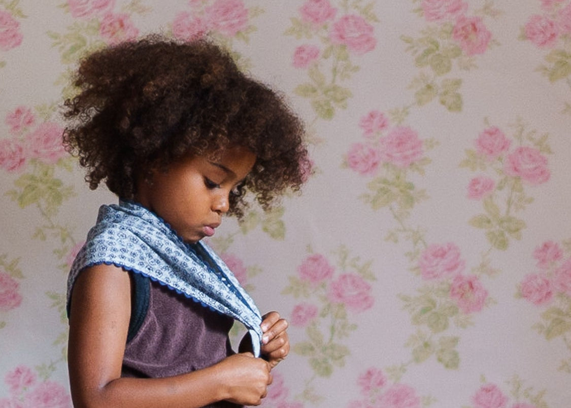 Young girl with curly hair wearing a soft, vintage-inspired blue floral scarf, showcasing natural, ethically made children’s clothing in a gentle, floral background.