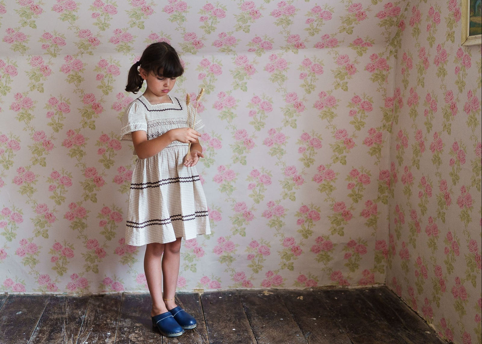A young girl wearing a vintage-style cream dress with smocked detailing, standing on a rustic wooden floor against a floral wallpapered wall, embodying classic and sustainable children's clothing.