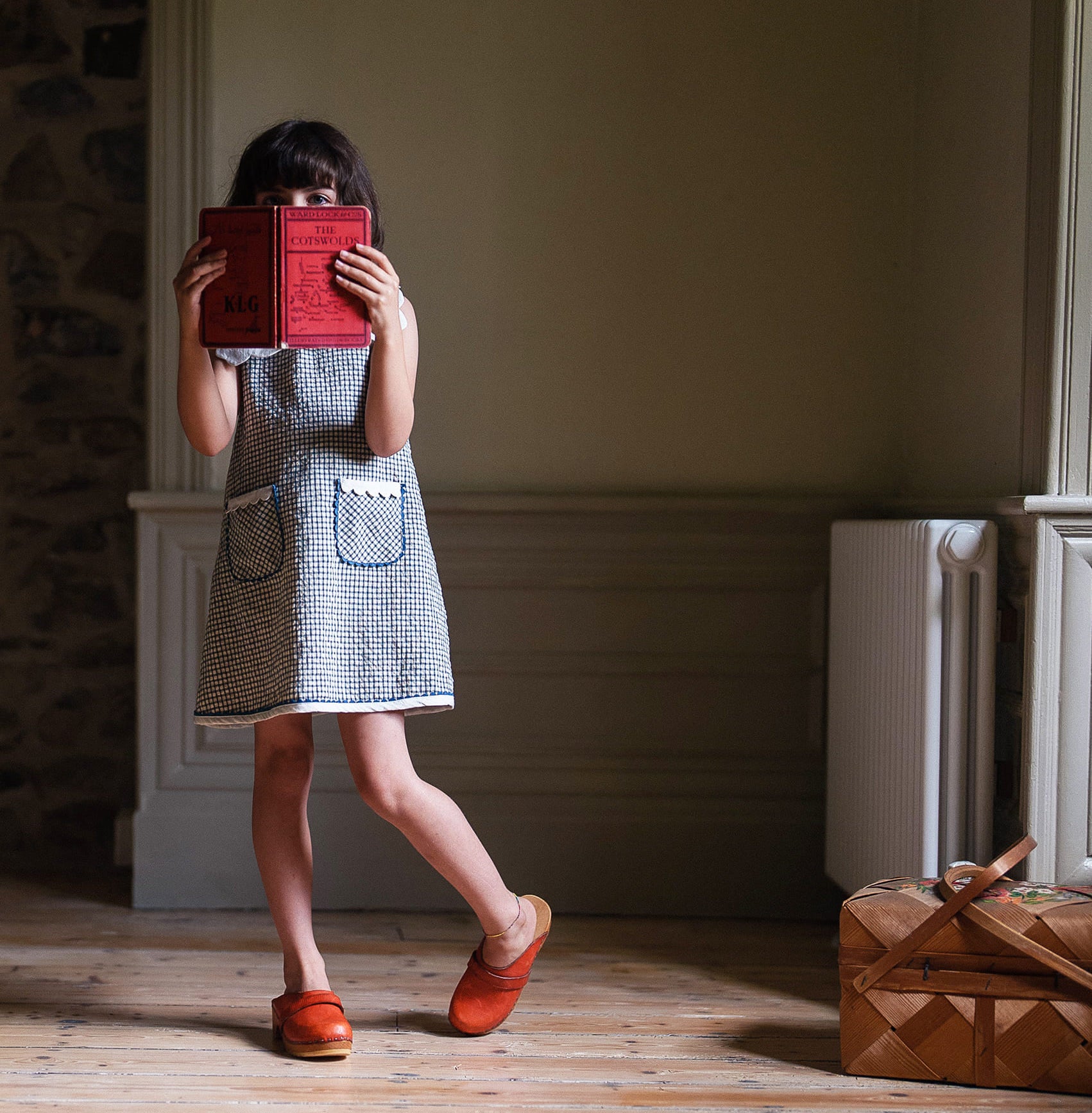 Young girl wearing a navy and white checkered cotton dress with front pockets, holding a red book in front of her face in a softly lit vintage-style room with wooden floors.