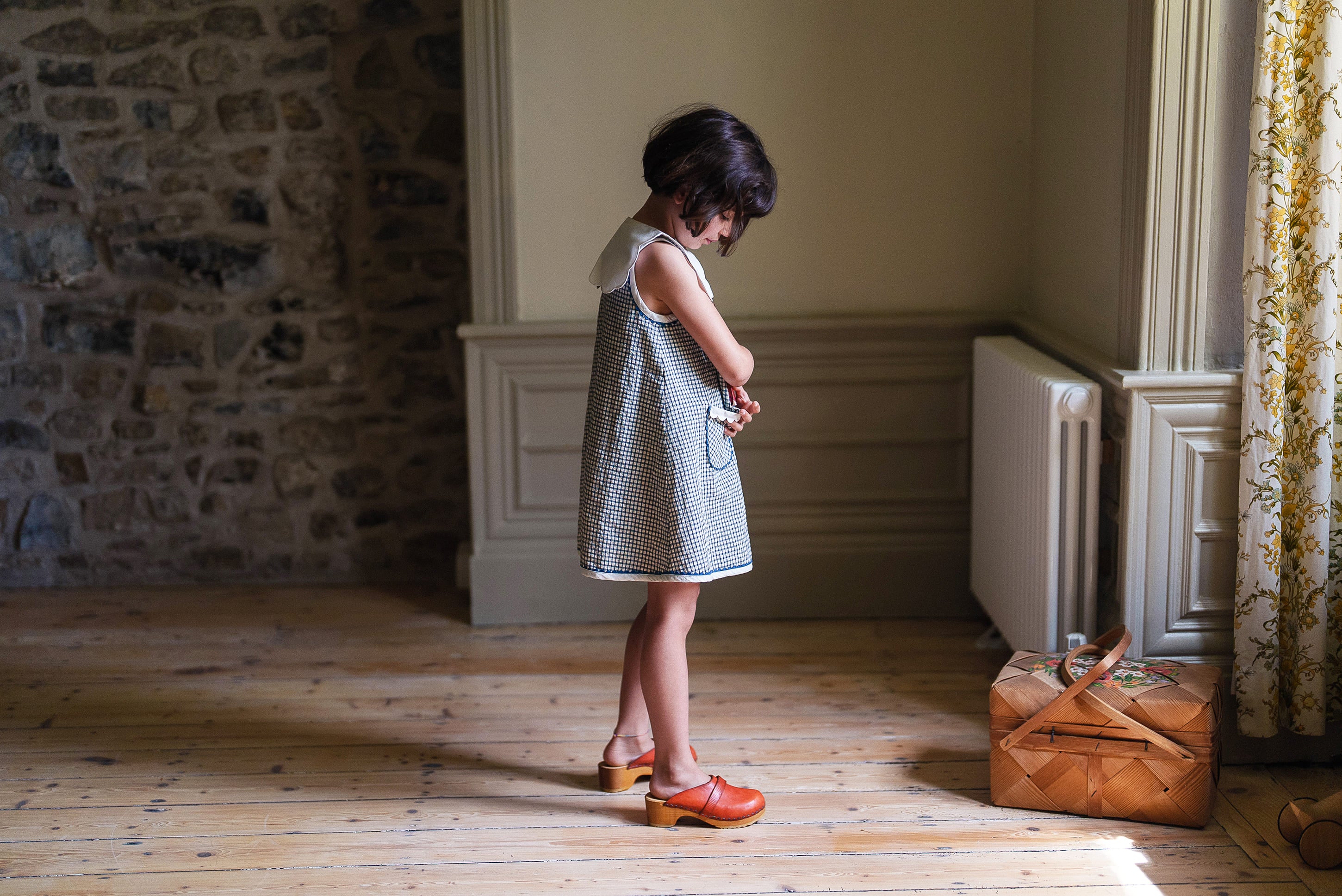Young girl wearing a navy and white checkered organic cotton dress standing in a vintage-style room with wooden floors, natural light, and a woven basket nearby.