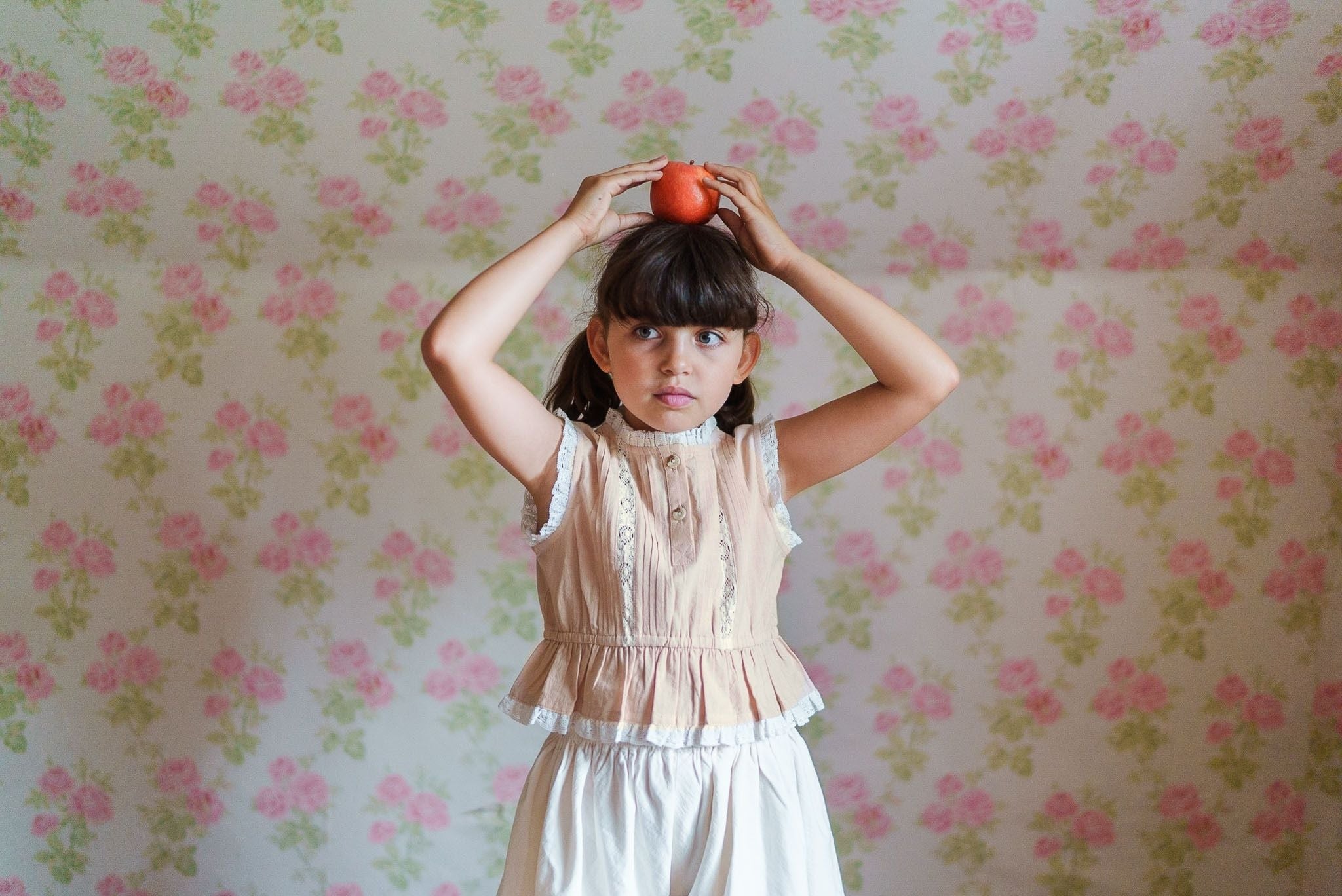 Young girl wearing a vintage style powder pink blouse made of organic cotton, holding an apple above her head against a floral wallpaper background.