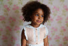 Young girl wearing a powder pink romper made from organic cotton, featuring vintage-inspired lace details and buttons, set against a floral background in a natural, ethical children’s clothing style.