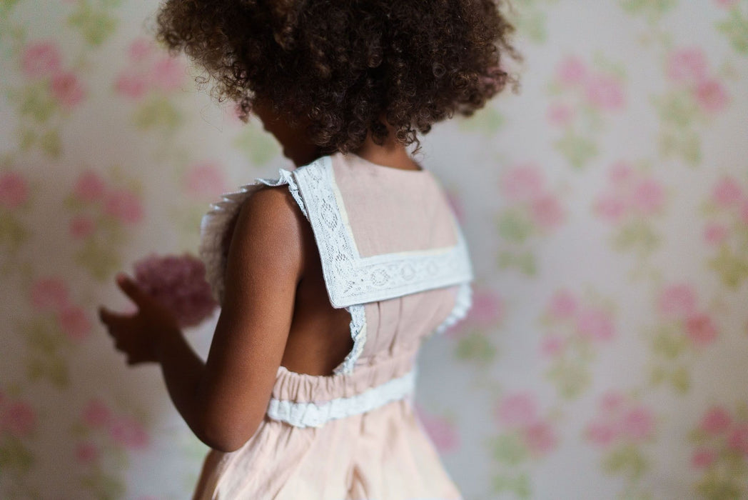 Young child wearing a vintage-style pink organic cotton romper with lace details, standing against a soft floral background, embodying slow, natural, and ethical children's clothing.