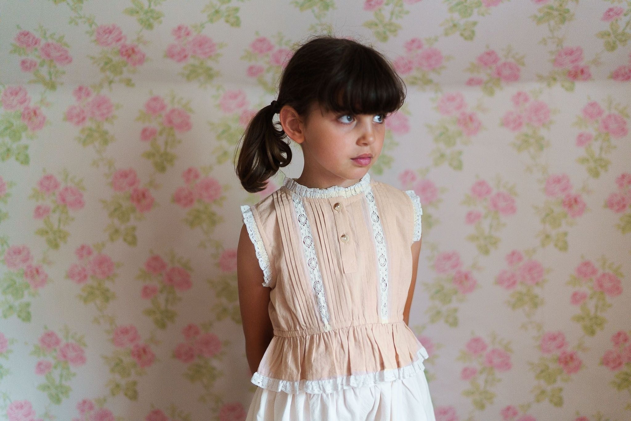 Young girl wearing a vintage style girls blouse in powder pink organic cotton with lace trim, standing against a floral wallpaper background in a softly lit room.