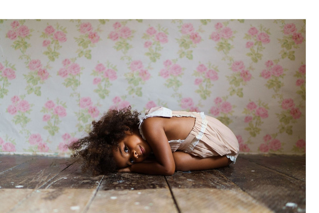 A young child lying on a wooden floor wearing a powder pink romper made from organic cotton, set against a vintage floral wallpaper backdrop, embodying slow, natural, and ethical children's clothing.
