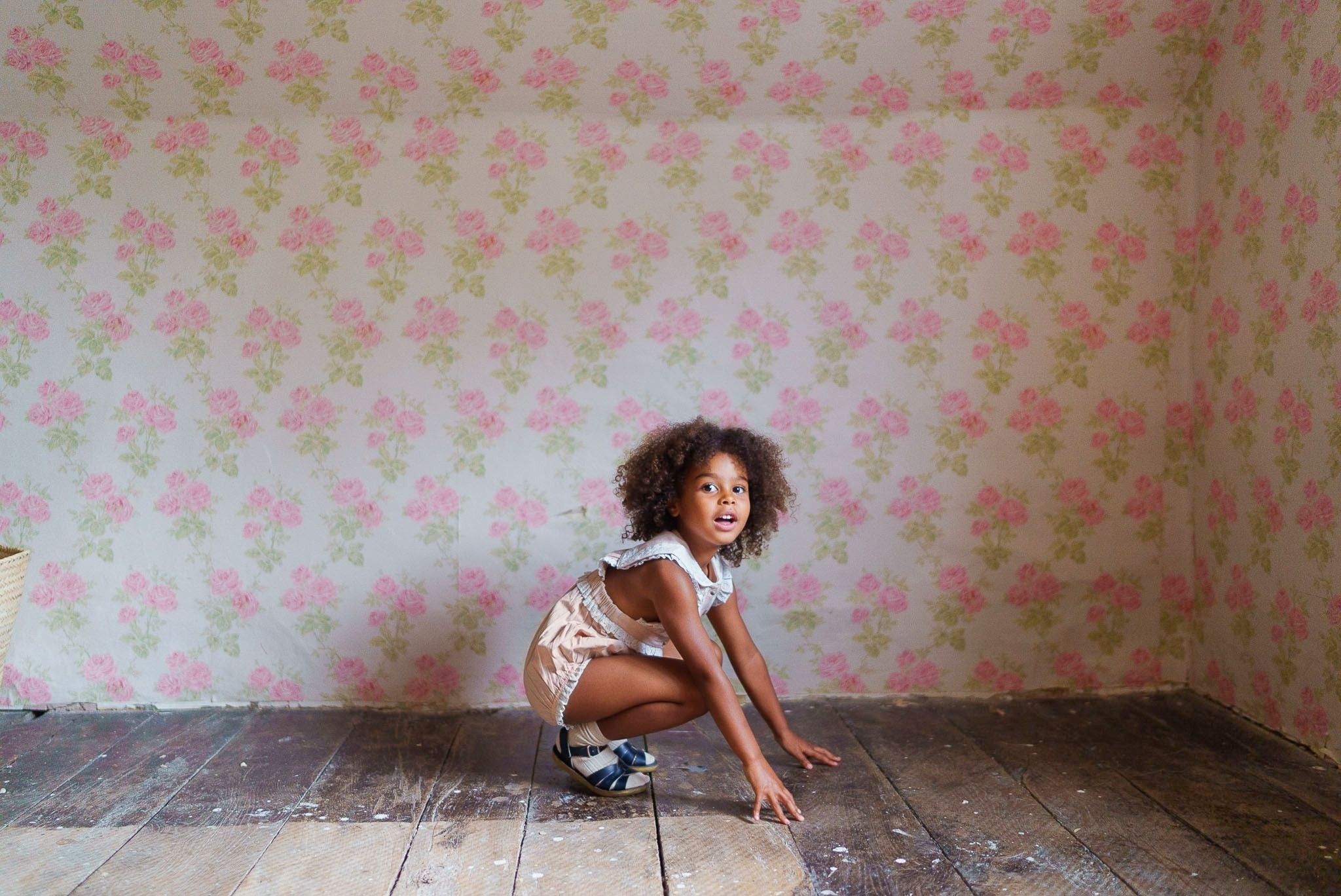 Young girl wearing a powder pink organic cotton romper with vintage style, playing on a wooden floor against a floral wallpaper backdrop in a natural, ethical children’s clothing setting.