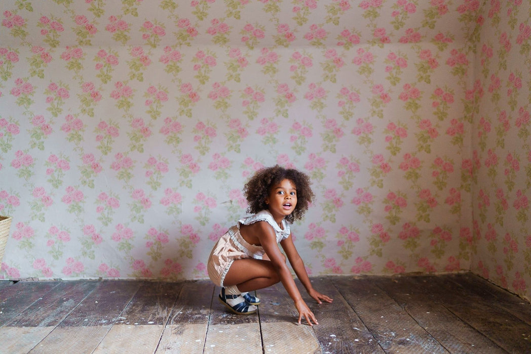 Young girl wearing a powder pink organic cotton romper with vintage style, playing on a wooden floor against a floral wallpaper backdrop in a natural, ethical children’s clothing setting.