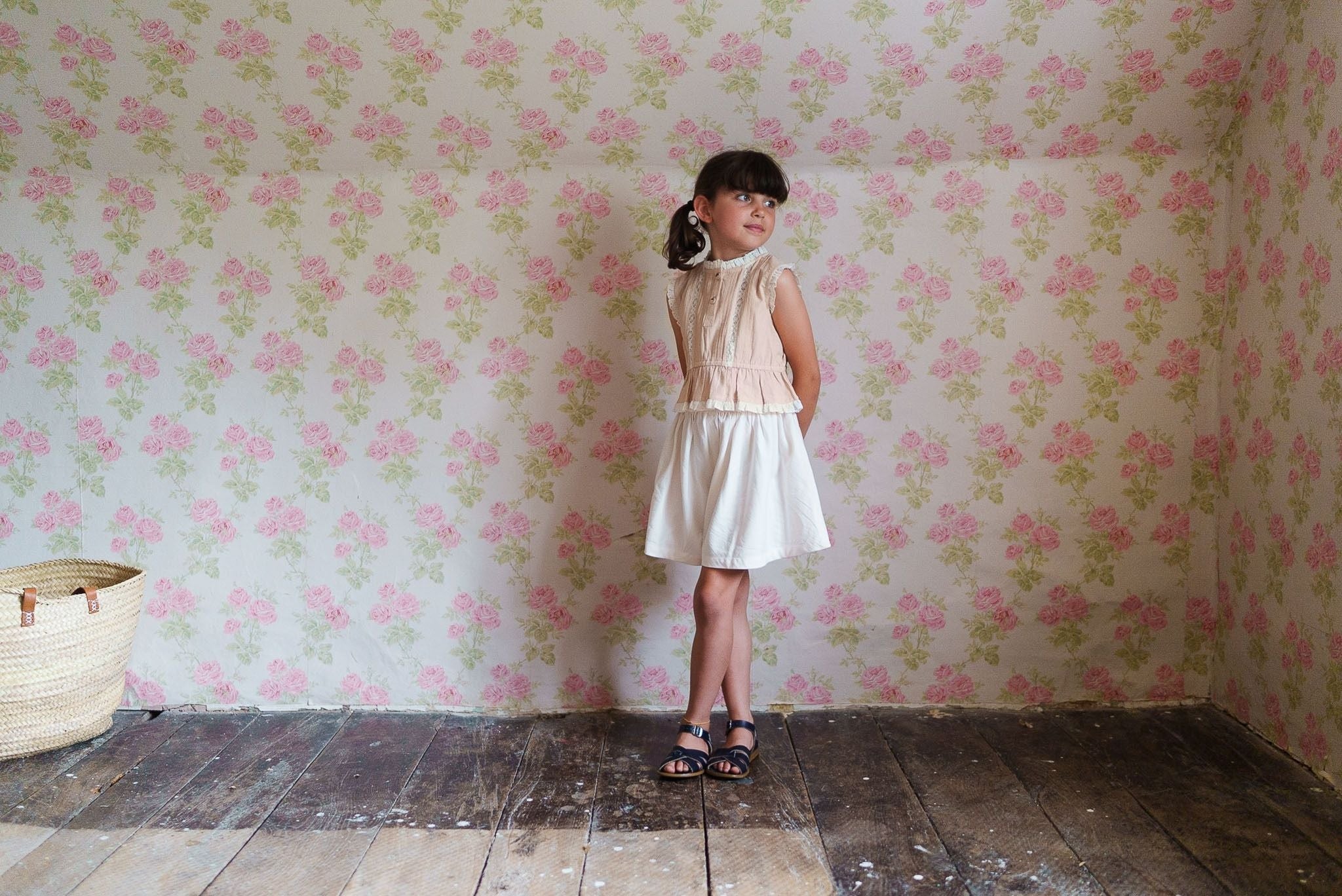 Young girl wearing a powder pink organic cotton blouse with a white skirt, standing in a vintage-style room with floral wallpaper and wooden floorboards.