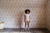 Young girl wearing a powder pink organic cotton romper with vintage-inspired details, standing in a floral wallpapered room showcasing sustainable, ethical children's clothing.