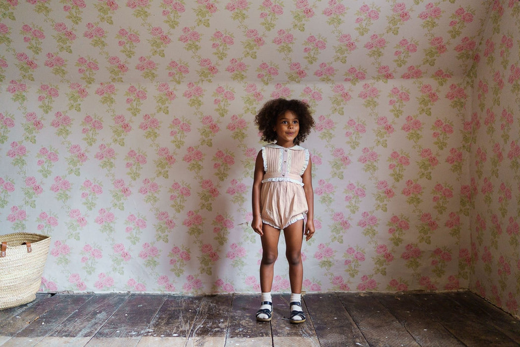 Young girl wearing a powder pink organic cotton romper with vintage-inspired details, standing in a floral wallpapered room showcasing sustainable, ethical children's clothing.