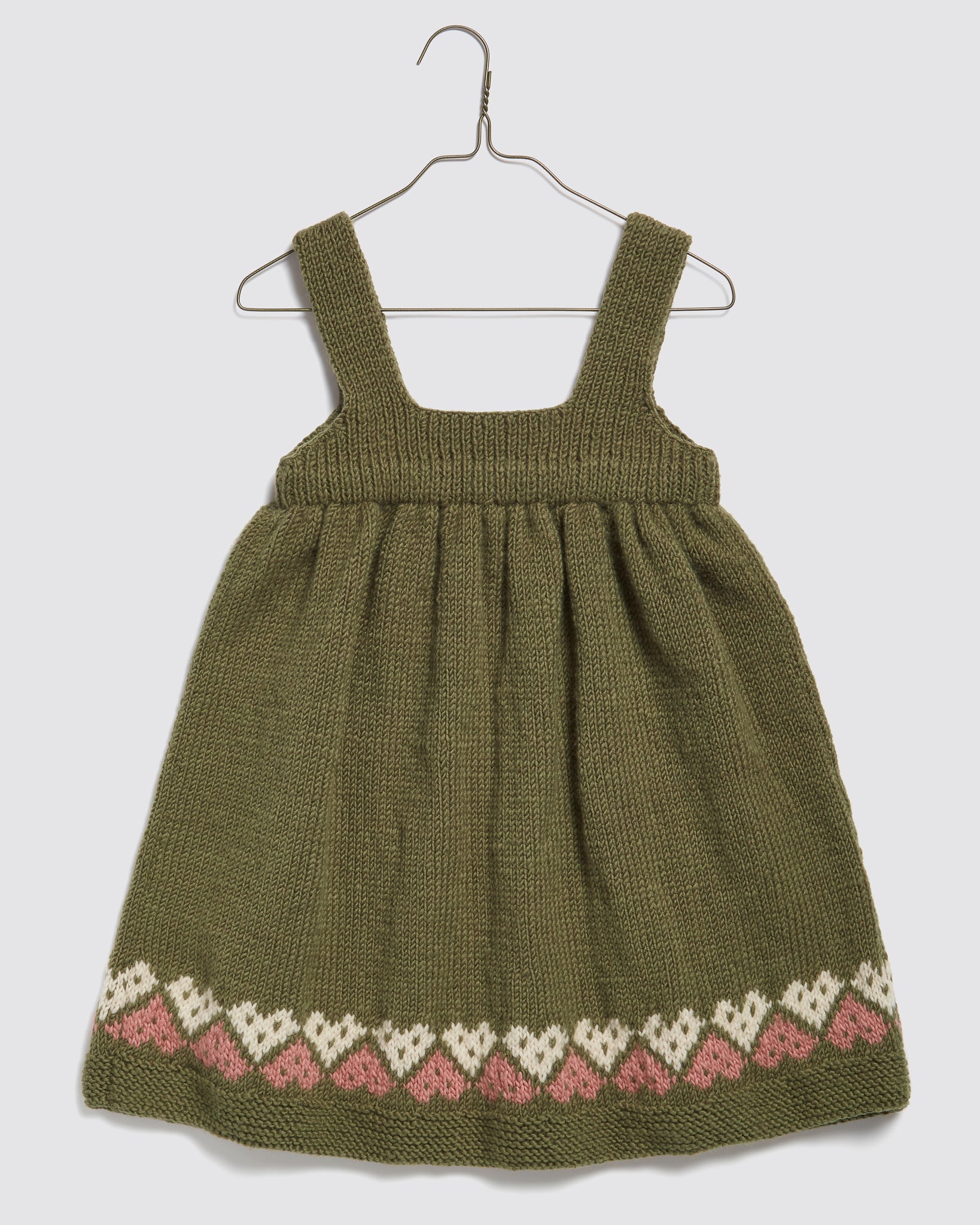 Hand-knitted olive green pinafore dress for kids made from soft merino wool, featuring a vintage-inspired heart pattern in beige and pink along the hem, crafted sustainably.