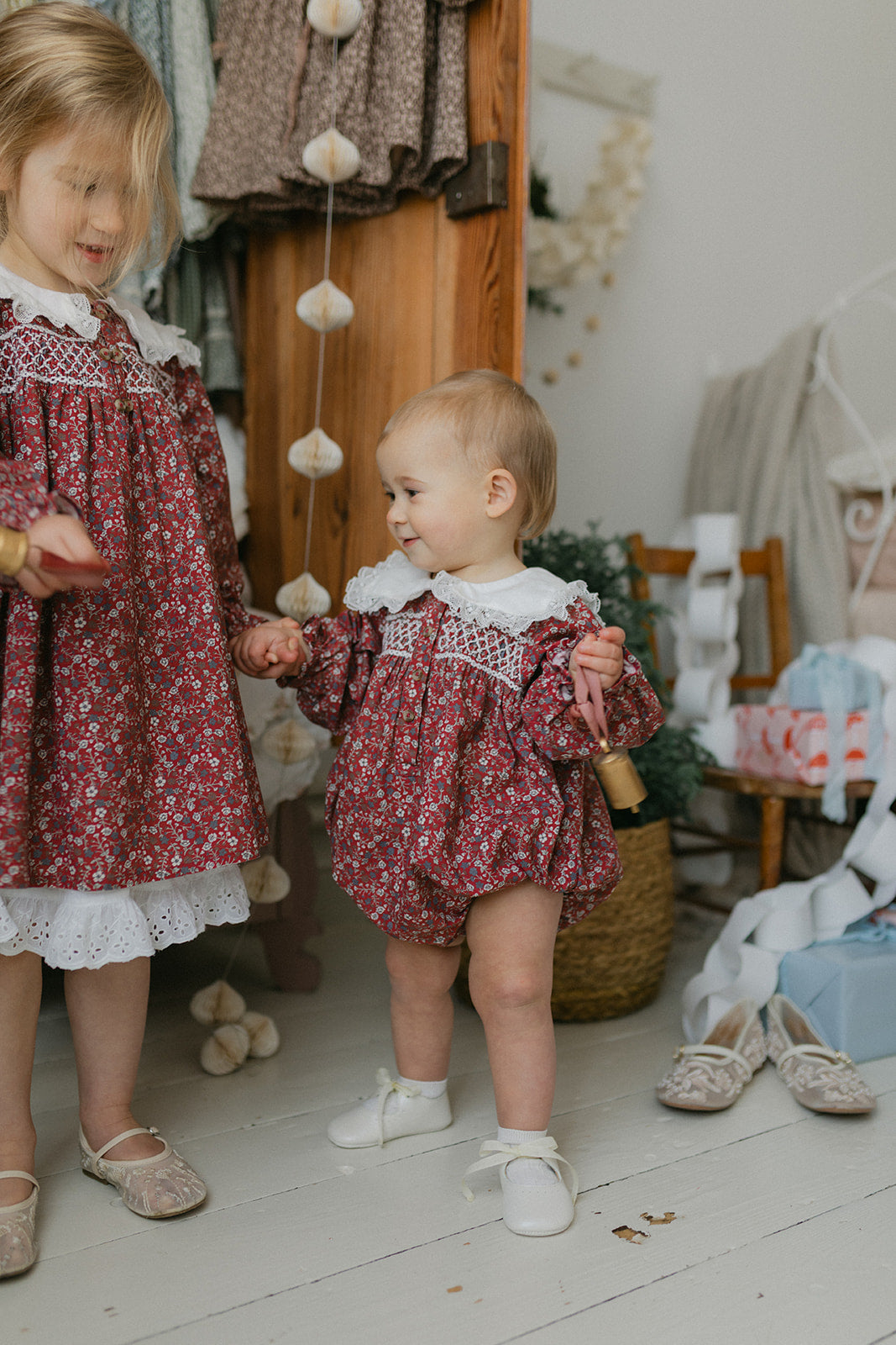 Two young children wearing vintage style, organic cotton floral rompers in warm clove tones, playing together in a cozy, sustainably styled room.