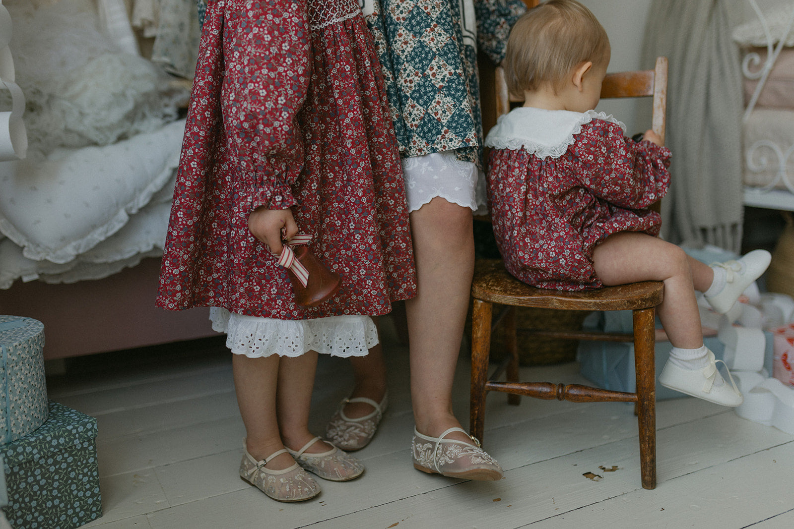Two children and an adult wearing vintage-style natural cotton clothing in warm floral patterns, featuring a baby in a floral romper sitting on a wooden chair in a cozy, softly lit room.