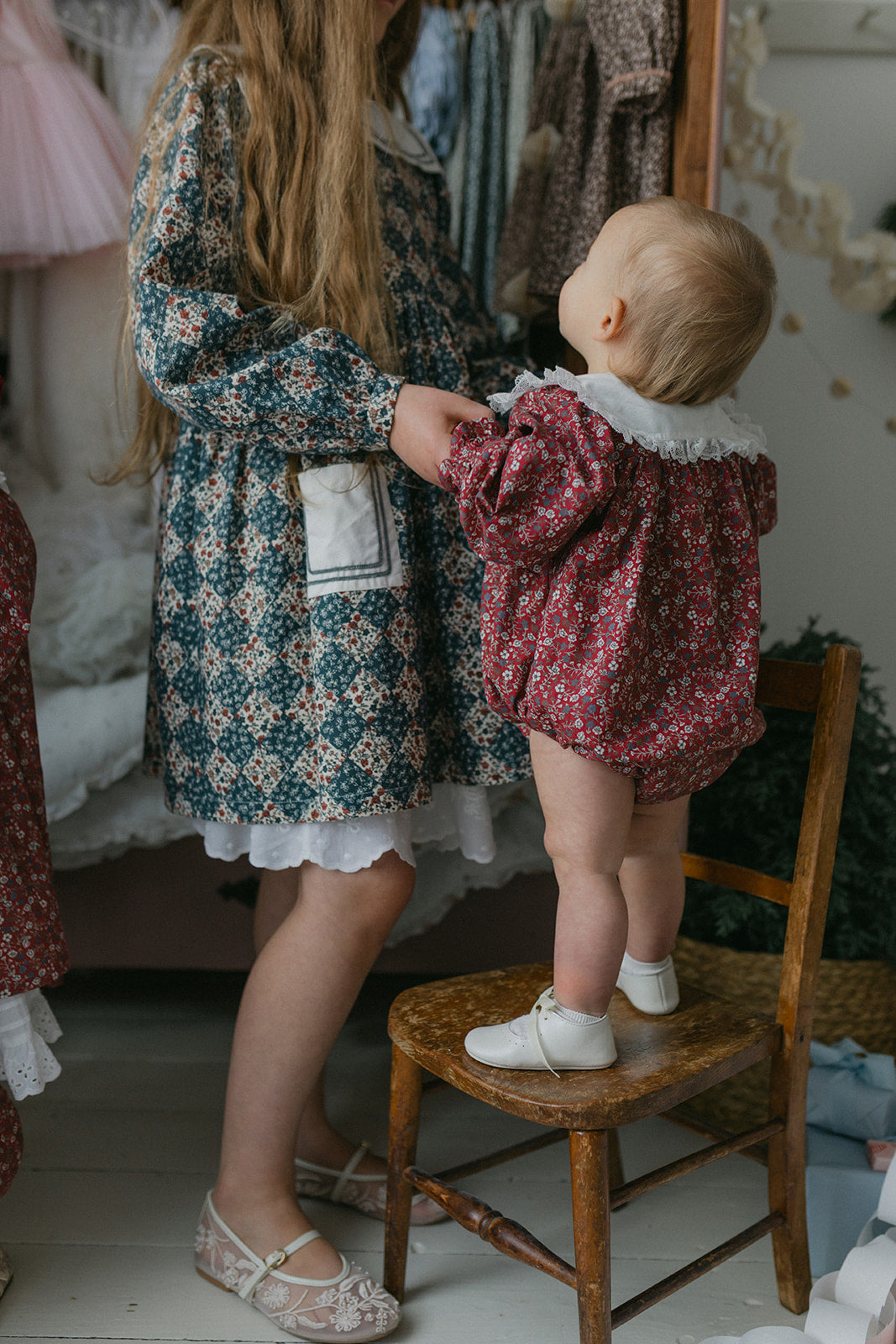 Toddler wearing a clove floral romper with lace collar standing on a wooden chair, holding hands with an older child in a vintage-style organic cotton dress, in a cozy room.