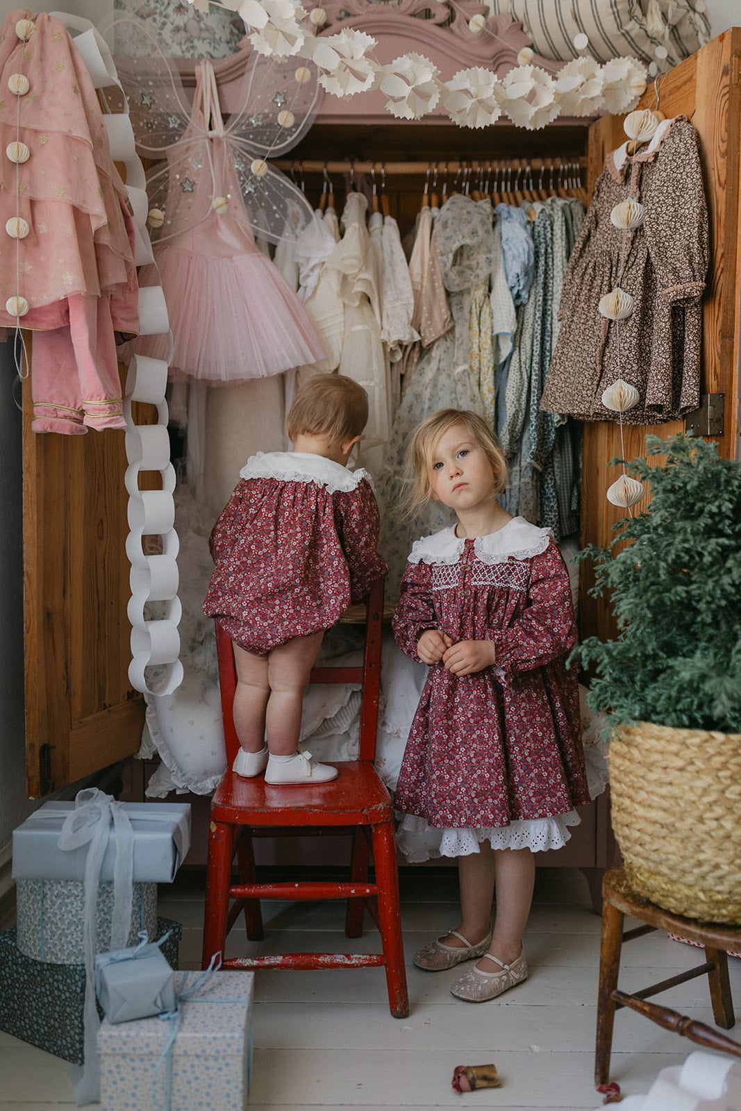 Two young children wearing vintage style clove floral rompers made from organic cotton stand in a cozy, softly lit room filled with traditional and sustainably made children's clothing.