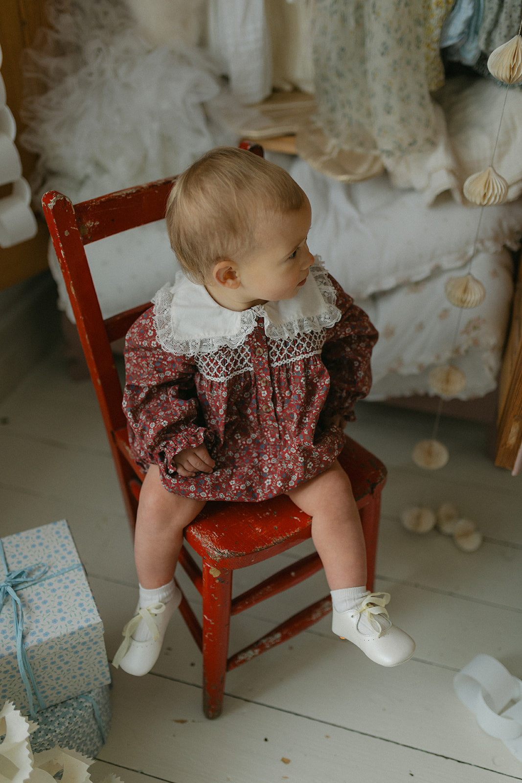 Baby wearing vintage style romper with delicate floral pattern and lace collar, sitting on a rustic red wooden chair in a cozy, natural setting.
