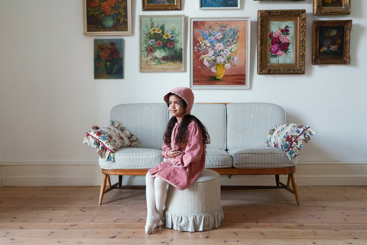 Young girl sat on pouffe wearing vintage-inspired soft pink cord bonnet and matching dress with cream tights, in front of striped sofa with classic flower paintings on the wall, showing vintage inspired and sustainable values.