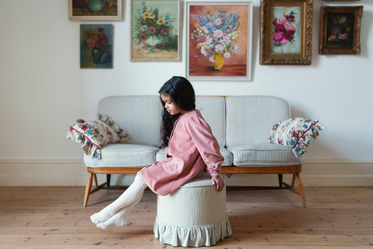 Young girl sat on pouffe wearing soft pink cord dress with long sleeves and contrasting pockets paired with cream tights in a vintage-inspired children's outfit.