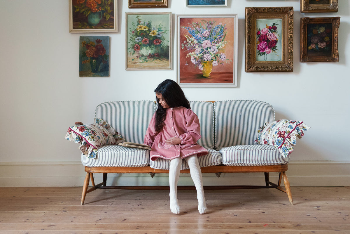 Young girl wearing soft pink cord vintage-inspired dress with cream tights sat on striped sofa with floral cushions and classic flower paintings on wall behind.