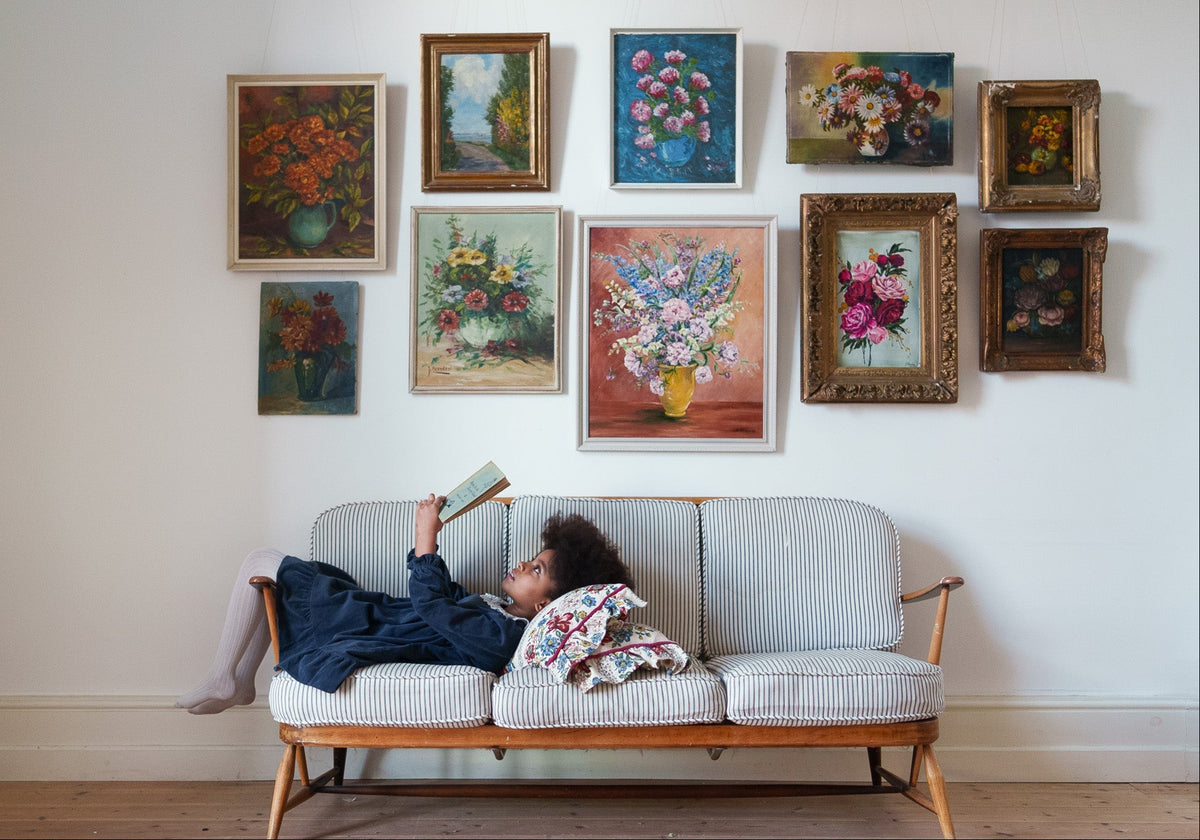 Child wearing a midnight navy velvet girls dress while reclining on a striped vintage sofa beneath an arrangement of floral paintings in a cosy room